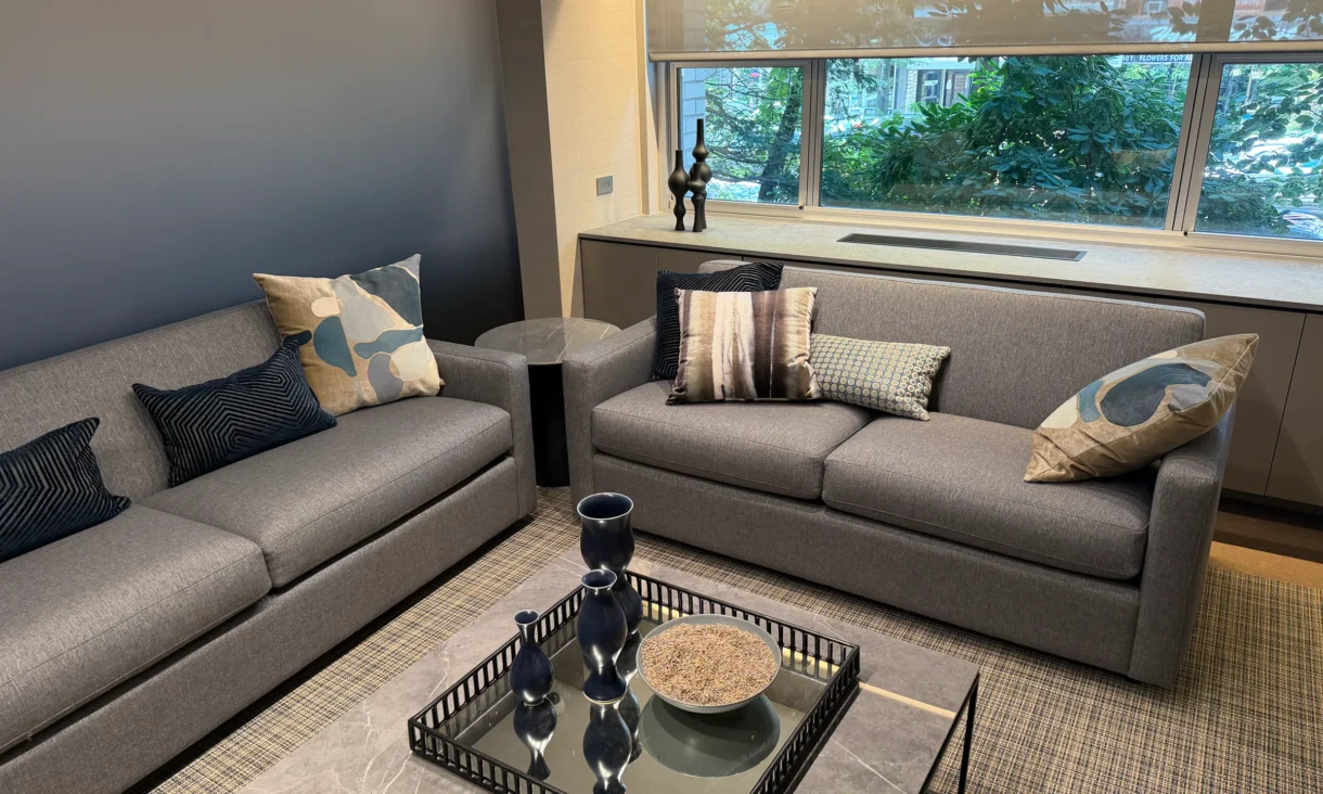 Cambridge Lounge seating area with gray sofas, patterned cushions, and a glass coffee table beside a window.
