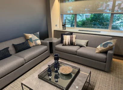 Cambridge Lounge seating area with gray sofas, patterned cushions, and a glass coffee table beside a window.